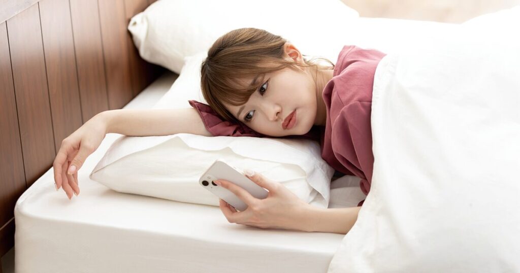 仕事のやる気がでず、朝ベッドの中でスマホを持ち、職場へ欠勤の連絡を入れるかどうか迷っている女性